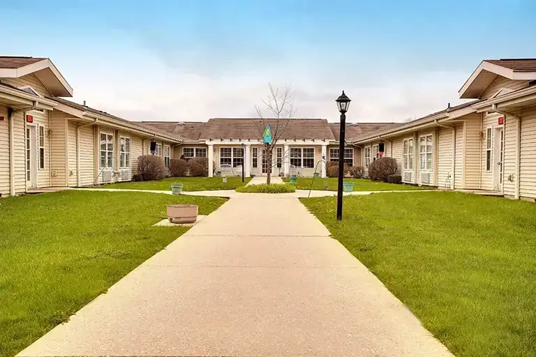 A wide sidewalk leads through a grassy courtyard between two single-story beige buildings with large windows under a blue sky, creating a welcoming atmosphere at Trustwell Senior Living. A wide sidewalk leads through a grassy courtyard between two single-story beige buildings with large windows under a blue sky, creating a welcoming atmosphere at Trustwell Senior Living.