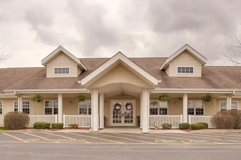Single-story beige Trustwell Senior Living building with a covered entrance, white columns, front porch, and decorative wreaths on double doors; parking spaces and cloudy sky in the foreground.