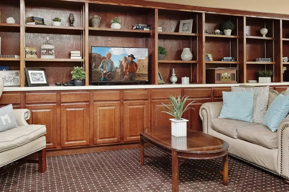 Living room at Trustwell Senior Living with wooden shelves, a TV showing two people outdoors, beige armchairs, a sofa with blue pillows, a coffee table, and decorative plants.