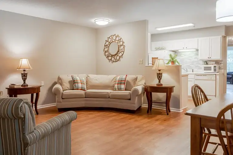 A cozy living room at Trustwell Senior Living features a beige sofa, two side tables with lamps, a striped armchair, and a dining table, all adjacent to a bright white kitchen. A cozy living room at Trustwell Senior Living features a beige sofa, two side tables with lamps, a striped armchair, and a dining table, all adjacent to a bright white kitchen.