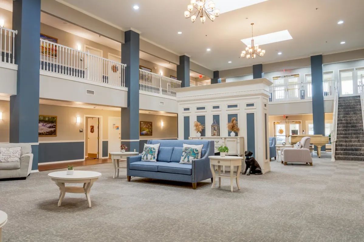 Spacious, well-lit lobby at Trustwell Senior Living with blue and white decor, comfortable sofas, coffee tables, sparkling chandeliers, and a staircase leading to a second-floor balcony.