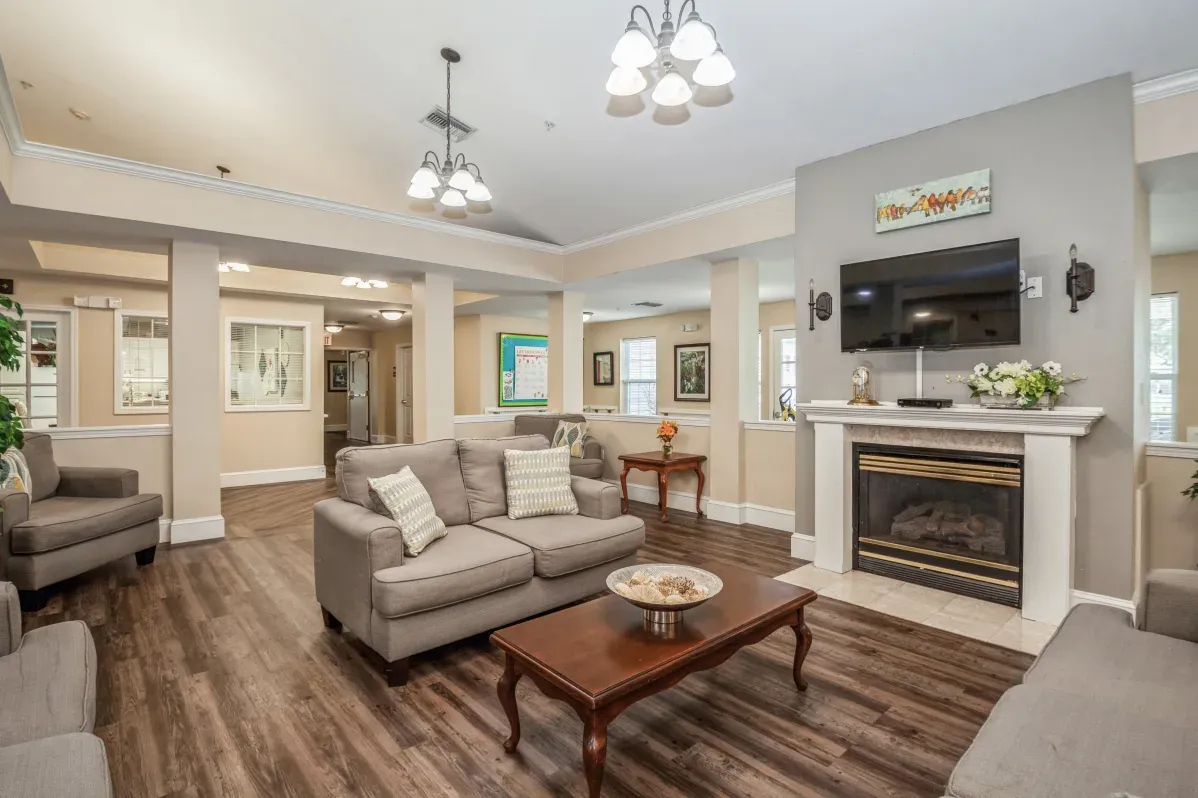A living room at Trustwell Senior Living with gray sofas, a wooden coffee table, a fireplace with a TV above it, and wood-style flooring under ceiling lights.
