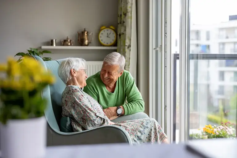 An elderly couple sits together in a bright Trustwell Senior Living room by a window, smiling and talking, with a yellow clock and potted plants in the background.
