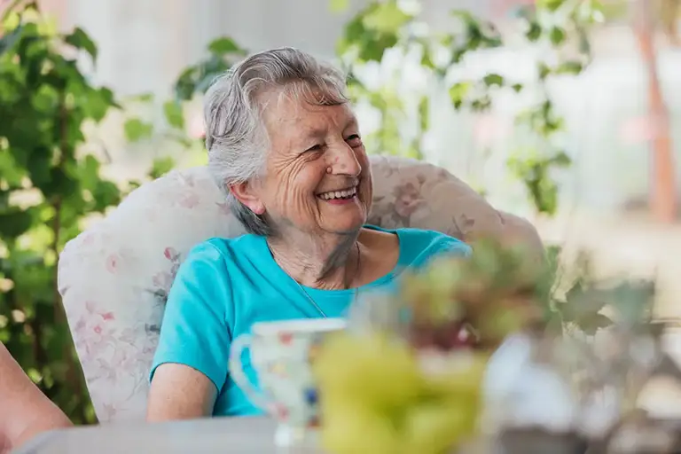 An elderly woman with gray hair and a blue shirt sits indoors at Trustwell Senior Living, smiling, with plants and a teacup in the background.