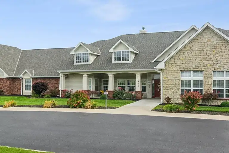A single-story Trustwell Senior Living building with stone and brick exterior, gabled roof, dormer windows, and a covered front entrance, surrounded by landscaping and a paved driveway. A single-story Trustwell Senior Living building with stone and brick exterior, gabled roof, dormer windows, and a covered front entrance, surrounded by landscaping and a paved driveway.